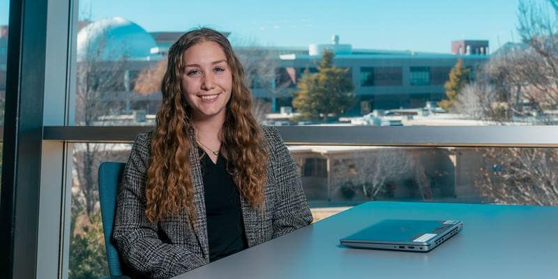 Meredith, a woman with light skin tone and long dark blond hair, sits at a table near a large window overlooking the Prescott Campus.